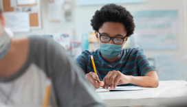 boy wearing a mask sitting at desk writing on a piece of paper