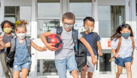 children wearing masks running out of a building with a soccer ball and backpacks